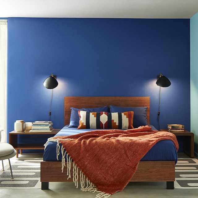 A contemporary bedroom with a dark blue painted wall, a light blue painted wall, a white ceiling, and a wooden bedframe and rust-coloured chenille throw.