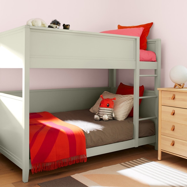 A charming children’s bedroom with a soft green-painted bunk bed, light pink walls, a blonde wood dresser, and pops of orange and coral colored bedding accents.