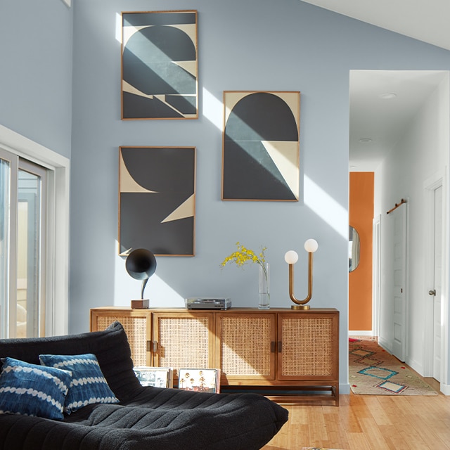 A light-filled living room with light blue-painted walls, a white ceiling, dark comfy sofa, three pieces of modern art over a cabinet and an orange accent wall at the end of a hallway.