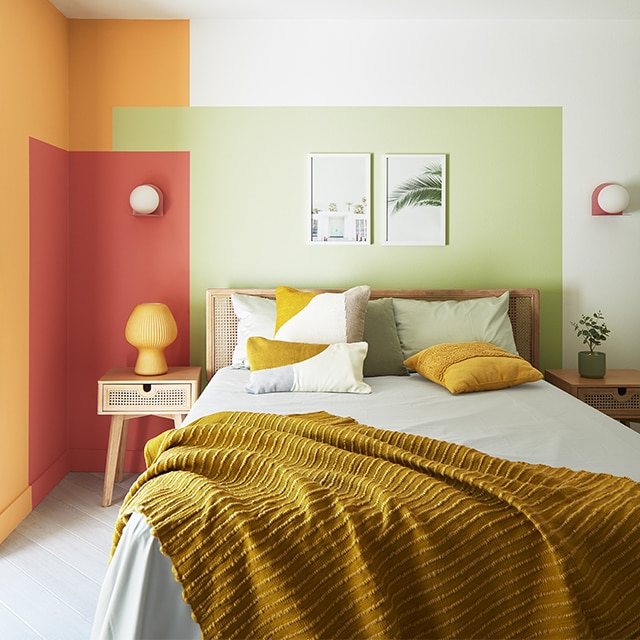 A small, contemporary bedroom with a painted colour block wall design in green, red and orange against white-painted walls, and a double bed with an orange throw.
