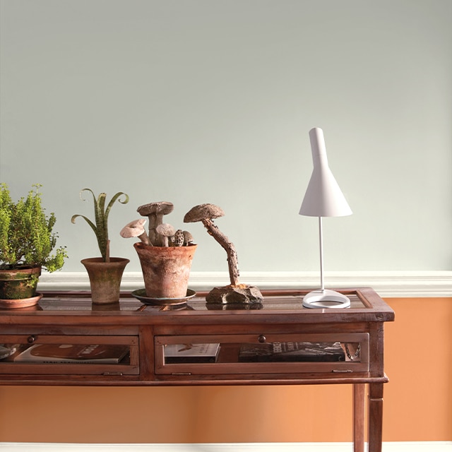 A split wall painted in cool gray and soft orange frames a wooden side table showcasing a white modern desk lamp and several planters.