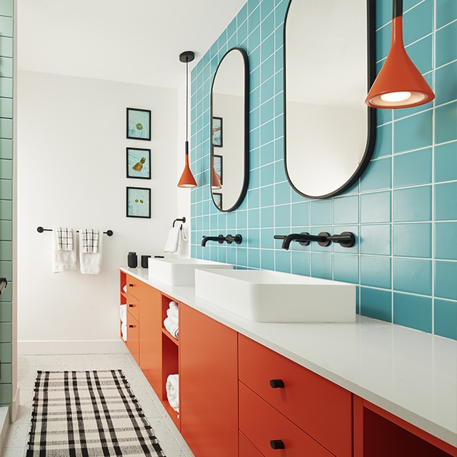A cheery bathroom with orange-red painted cabinetry, bright blue tile walls, red pendant lighting and white-painted walls and ceiling. 