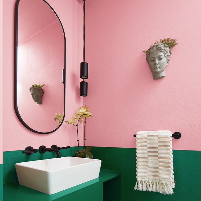 Un pequeño y divertido tocador con una pared dividida que presenta un rosa saturado en la parte superior, Verde Bosque en la inferior, un espejo ovalado y un lavabo blanco.