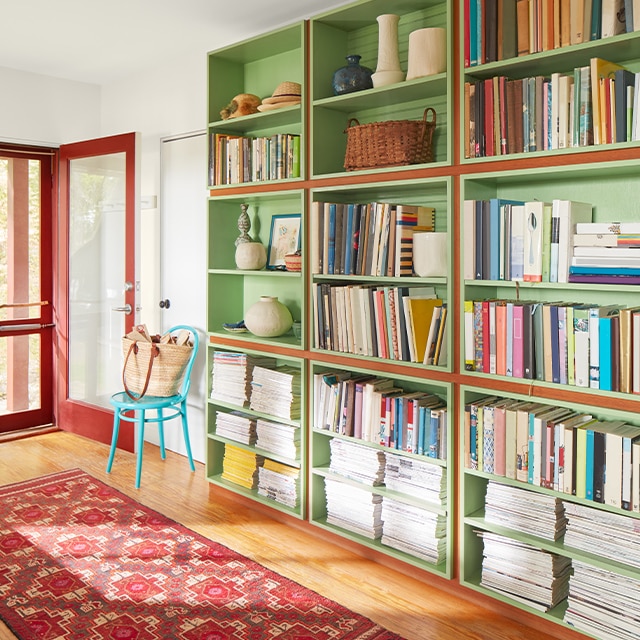 A sunlit entryway with green-painted bookshelves, wood floors, a red-painted front door, and a red patterned rug.