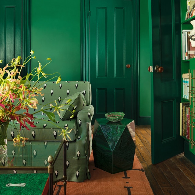 A green-painted living room with green-printed couch, stone end table, glass coffee table, and bookshelves matching the walls. 