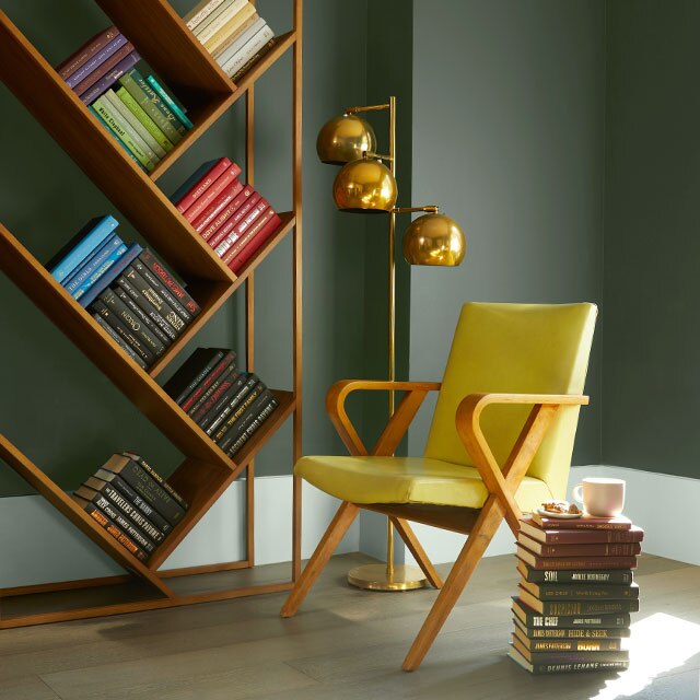 A home office with green-painted walls, green trim, brass bookshelf and lighting, and a stack of books next to a yellow chair.