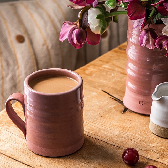 A muted brown ceramic mug and a vase of pink flowers atop a wooden table.