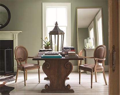 Table en bois foncé et deux chaises dans une pièce vert tendre, avec un miroir pleine longueur incliné et une grande fenêtre.