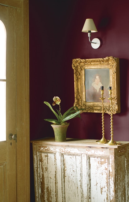 Cette entrée inondée de lumière met en vedette deux chandelles coniques et une plante fleurie en pot posées sur une table ancienne patinée par le temps; en arrière-plan des murs de couleur prune foncée.