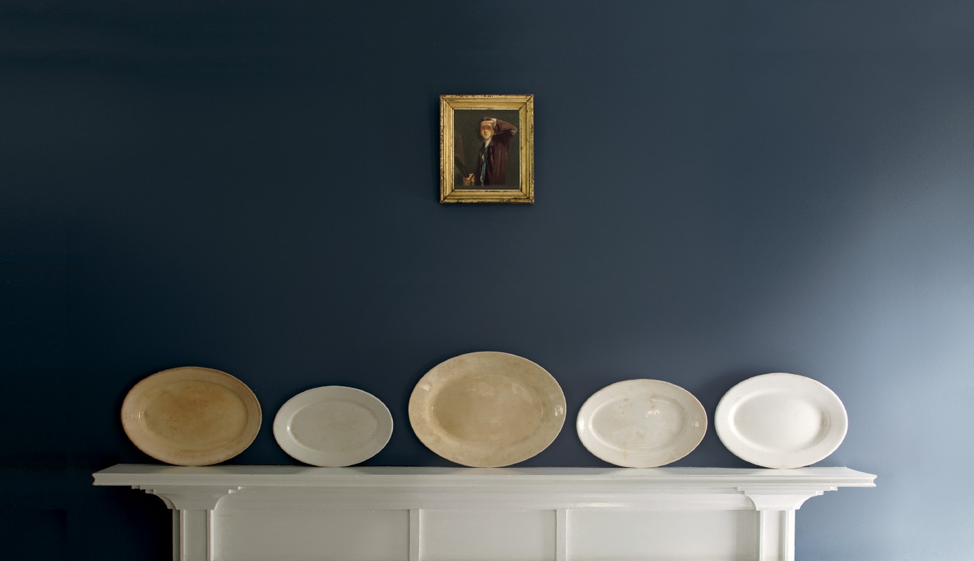 A white wood mantel features five decorative plates of varying sizes in neutral tones horizontally placed against a blue wall; a small antique painting hangs above.