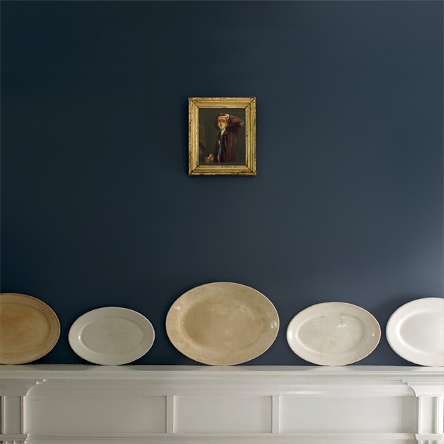 Una repisa de la chimenea de madera blanca presenta cinco platos decorativos de diferentes tamaños en tonos neutros colocados horizontalmente contra una pared azul; un pequeño cuadro antiguo está colgado arriba.