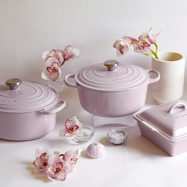 An array of Le Creuset cookware in Shallot, the newest Le Creuset color, a lovely lavender-gray, alongside several fresh flowers set off from off-white wall.