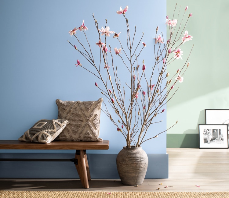 Un coin-salon deux tons arborant un mur bleu à l’avant-plan et un autre vert, un banc en bois, des branches dans un vase et des tableaux posés contre le mur du fond. 