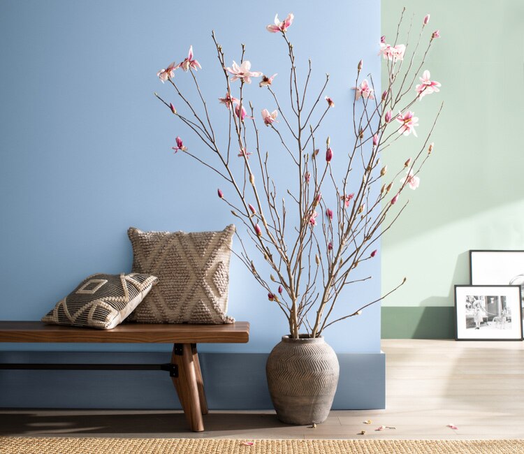 Two-toned sitting area with one blue wall in front and one green, wooden bench, vase with branches, and paintings propped against the back wall. 