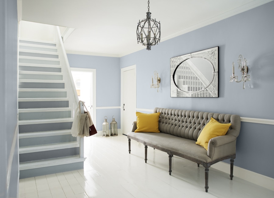 Blue-gray living area with two-toned stairs, wainscoting, gray couch with yellow pillows, small cage chandelier, and wall art.