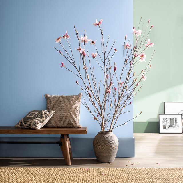 Two-toned sitting area with one blue wall in front and one green, wooden bench, vase with branches, and paintings propped against the back wall. 
