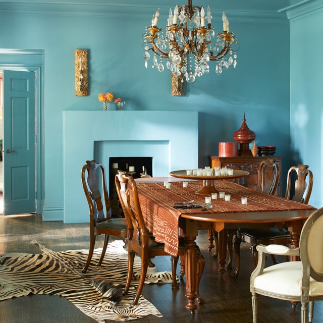 An opulent dining room with blue-painted walls, ceiling and fireplace, and a chandelier over a large wooden dining table and chairs. The style is classic with an exotic twist.