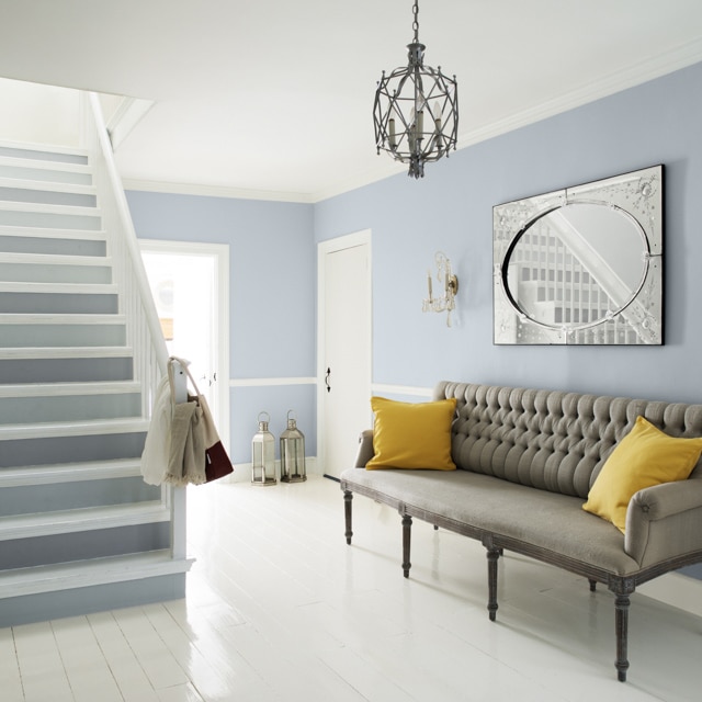 Blue-gray living area with two-toned stairs, wainscoting, gray couch with yellow pillows, small cage chandelier, and wall art.