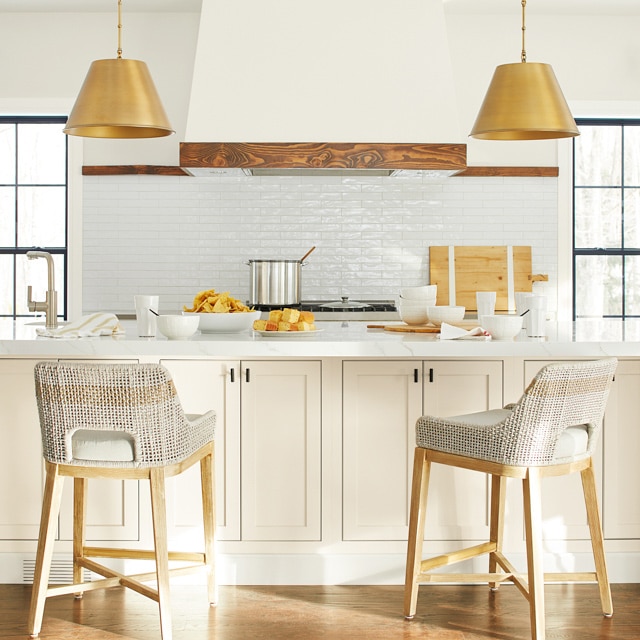 Kitchen with white walls and black trim, white island counters with beige stools, golden overhead lighting, and various kitchen utensils.