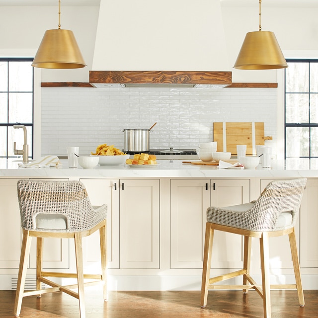 Cocina con paredes blancas y marcos negros, encimeras de isla blancas con banquetas beige, lámparas de techo doradas y diversos utensilios de cocina.