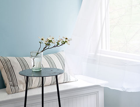 Light blue-painted walls with an open window and shear white curtains blowing in the wind featuring a bench with a striped pillow and black side table.