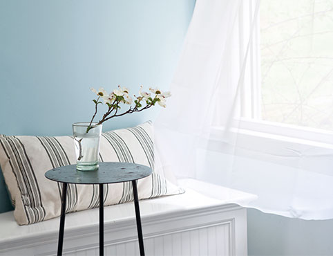 Light blue-painted walls with an open window and shear white curtains blowing in the wind featuring a bench with a striped pillow and black side table. Light blue-painted walls with an open window and shear white curtains blowing in the wind featuring a bench with a striped pillow and black side table.