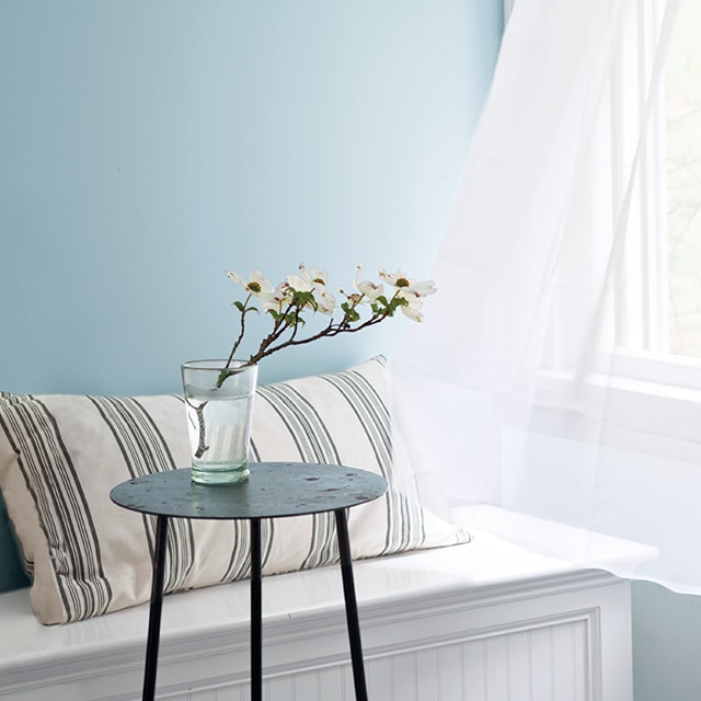 Light blue-painted walls with an open window and shear white curtains blowing in the wind featuring a bench with a striped pillow and black side table.