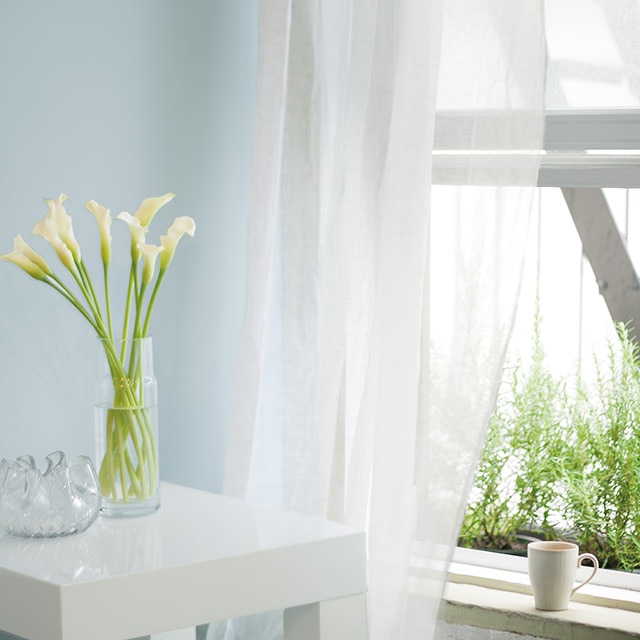 Un vase de fleurs est posé sur une petite table de chevet carrée près d’une fenêtre ouverte voilée d’un fin rideau blanc et d’un mur bleu pâle.