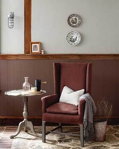 A wing chair and side table against deep red-wood paneled wainscoting, and light gray paint color on the upper wall.