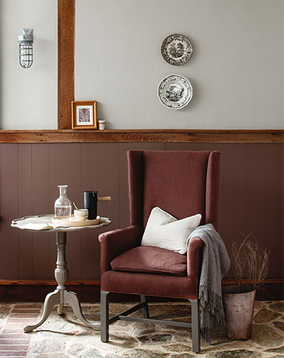 A wing chair and side table against deep red-wood paneled wainscoting, and light gray paint colour on the upper wall.