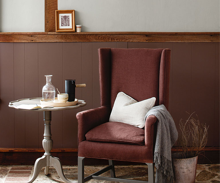 A wing chair and side table against deep red-wood paneled wainscoting, and light gray paint colour on the upper wall.