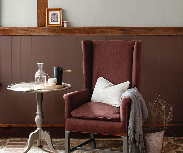 A wing chair and side table against deep red-wood paneled wainscoting, and light gray paint color on the upper wall.