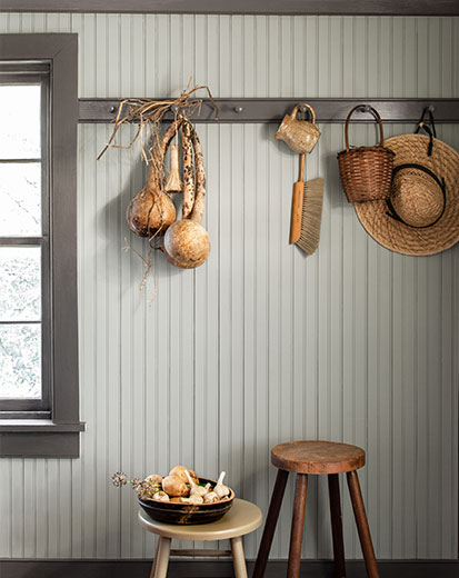 Un vestibule présentant un mur en lambris gris-blanc, deux tabourets, des chapeaux de paille et des paniers suspendus à une moulure gris foncé.