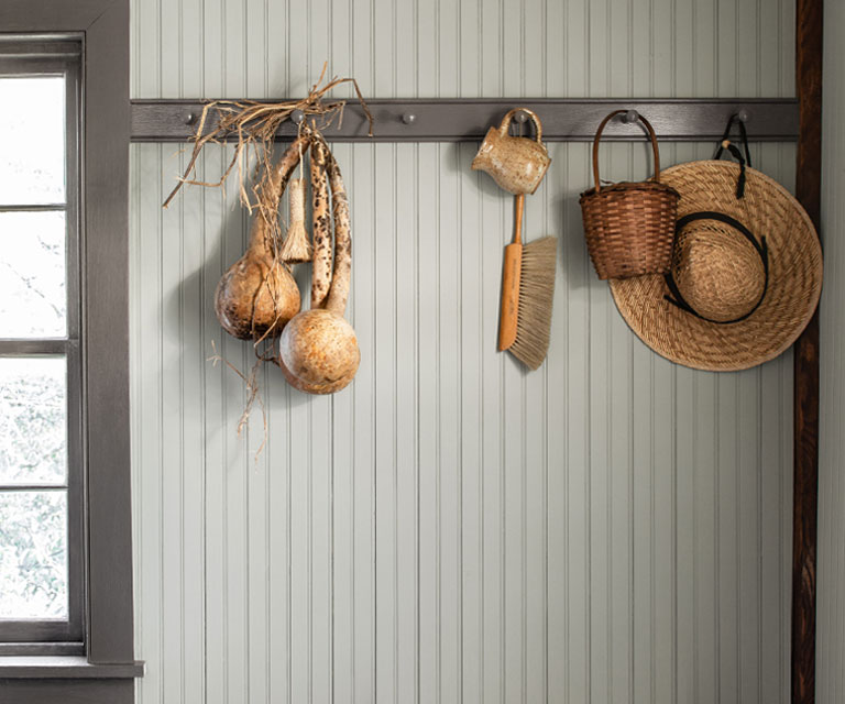 A gray-white paneled wall in an entryway with two stools; straw hats and baskets hang on dark gray trim and two stools.