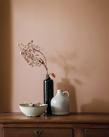 Un vase, une cruche et un bol posés sur une commode en bois contre un mur peint dans un brun doux teinté de rose.