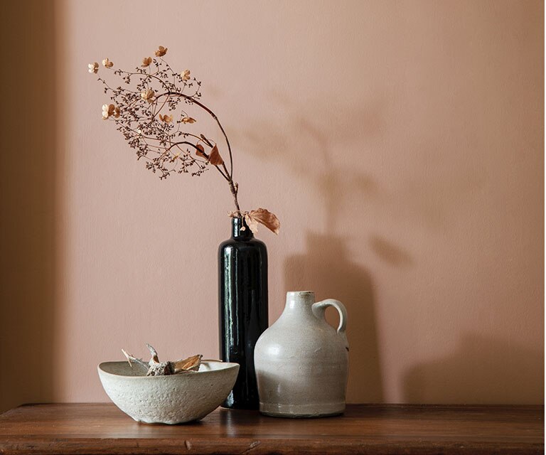 Un vase, une cruche et un bol posés sur une commode en bois contre un mur peint dans un brun doux teinté de rose.