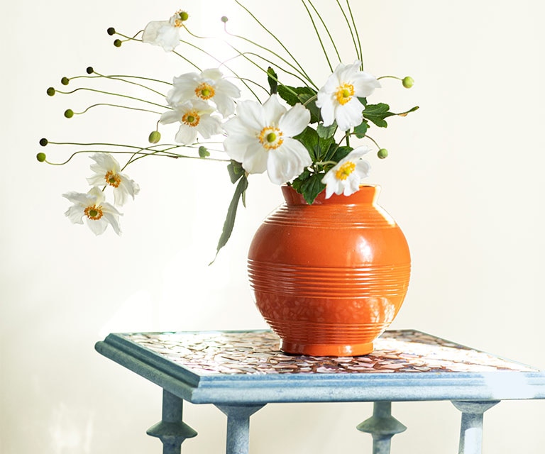 Une table bleue sur laquelle est posé un vase rempli de fleurs blanches contre un mur peint en blanc cassé.