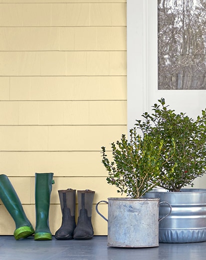 The front porch of a home with yellow siding, window in white trim, potted plants, and rubber boots.