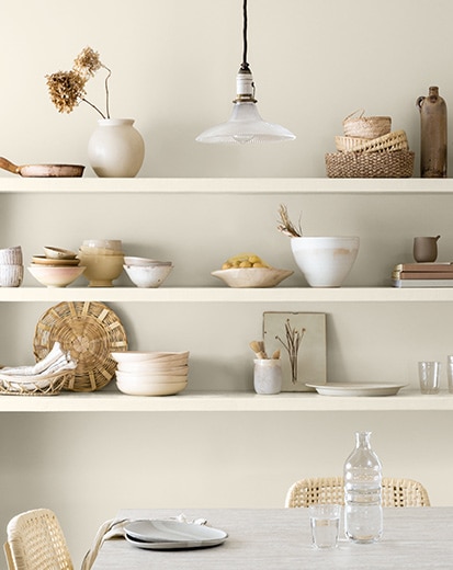 Une cuisine arborant des étagères ouvertes contenant des bols et des plats blancs; une table et des chaises blanches sur un mur peint en blanc.