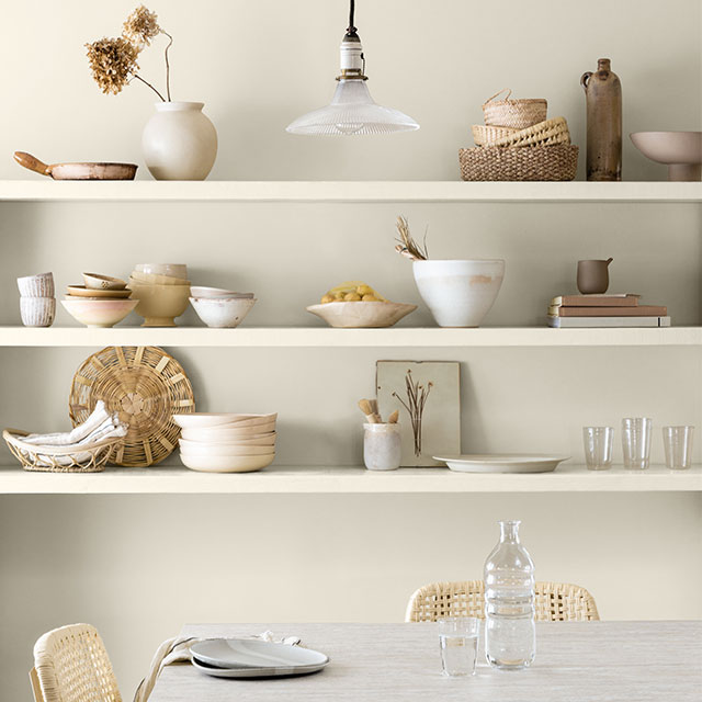 Une cuisine arborant des étagères ouvertes contenant des bols et des plats blancs; une table et des chaises blanches sur un mur peint en blanc.