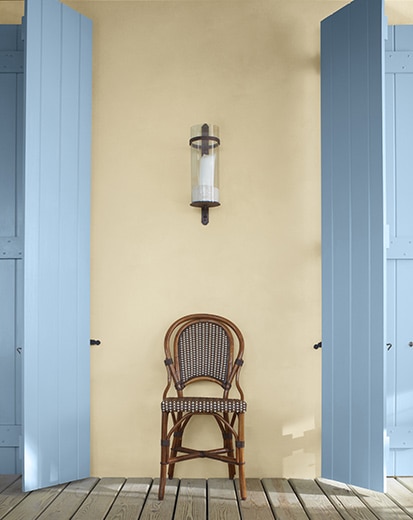 A porch featuring a chair between two light blue-painted shutters against a pale yellow-painted wall.