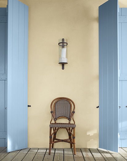 A porch featuring a chair between two light blue painted shutters against a pale yellow-painted wall.