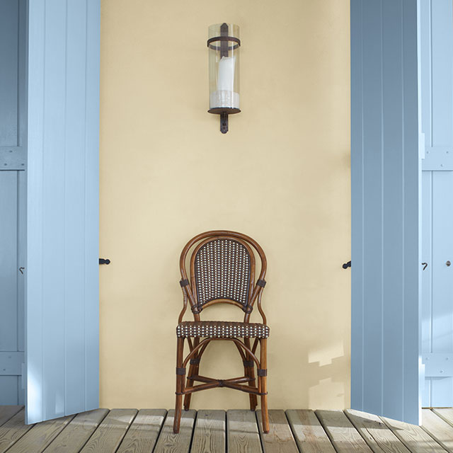 Un porche arborant une chaise entre deux volets peints en bleu clair sur un mur peint en jaune pâle.
