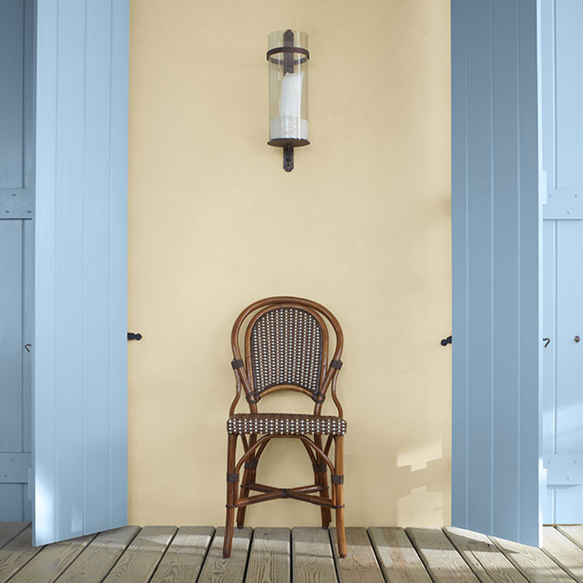 A porch featuring a chair between two light blue painted shutters against a pale yellow-painted wall.