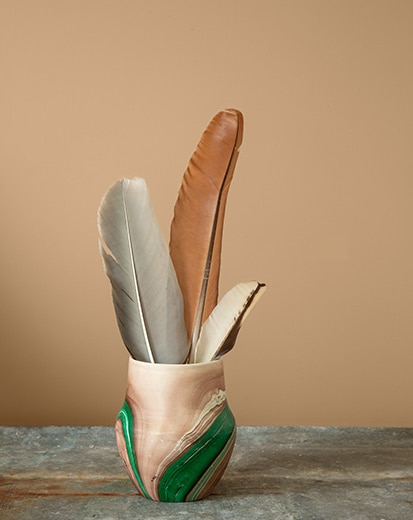 A green and cream-coloured vase holding feathers on a tabletop in front of a tan-painted wall.