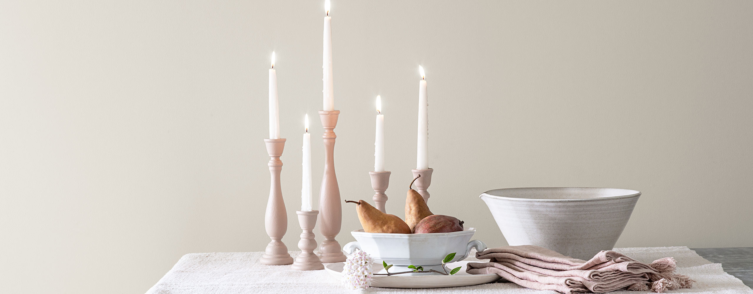 Lit tapered candles, two white porcelain bowls and off-white cloth napkins sit on a table against a white-painted wall.