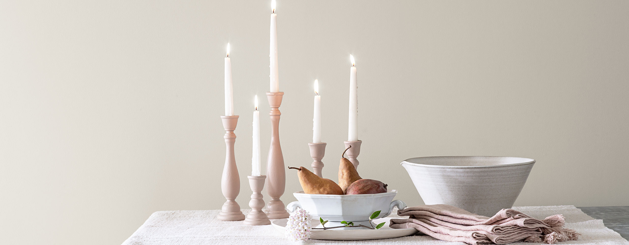 Lit tapered candles, 2 white porcelain bowls and off-white cloth napkins sit on a table against a white-painted wall.