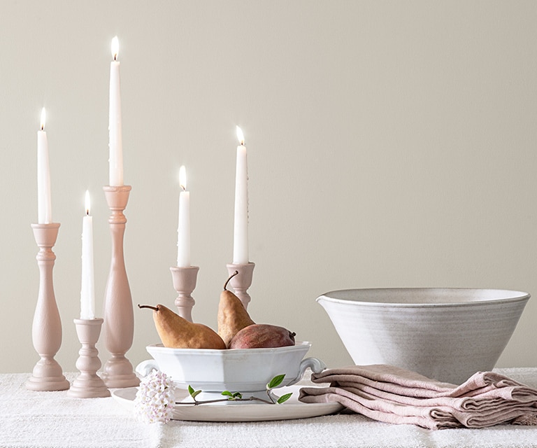 Lit tapered candles, two white porcelain bowls and off-white cloth napkins sit on a table against a white-painted wall.