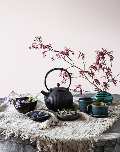A charcoal-gray cast iron tea pot, bowls, and a flowering tree stem against a gray-violet-painted wall.