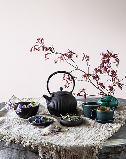 Une théière en fonte gris anthracite, des bols et une tige d’arbre en fleurs contre un mur peinturé en violet-gris.